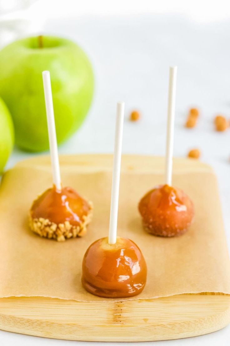 three caramel apples on a cutting board with two white sticks sticking out of them