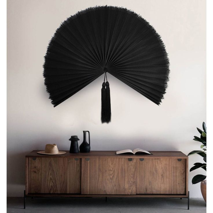 a large black fan hanging on the wall above a wooden cabinet with books and vases