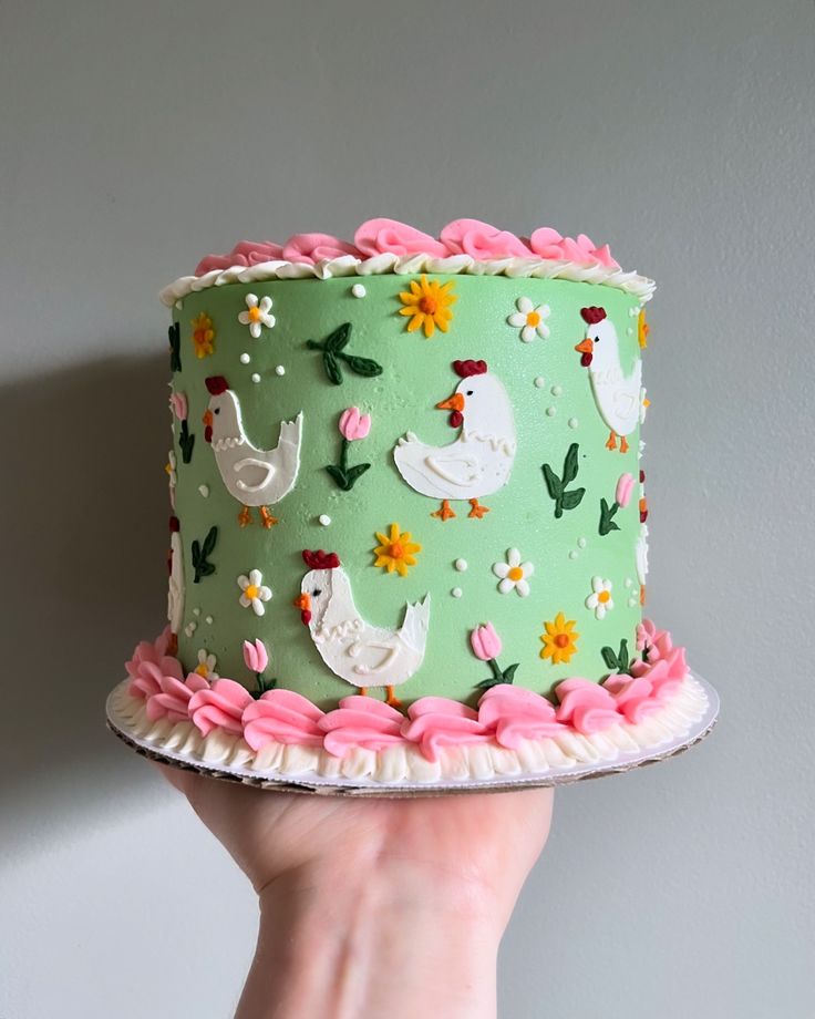 a hand holding a green cake with white chickens and pink flowers on it, in front of a gray wall