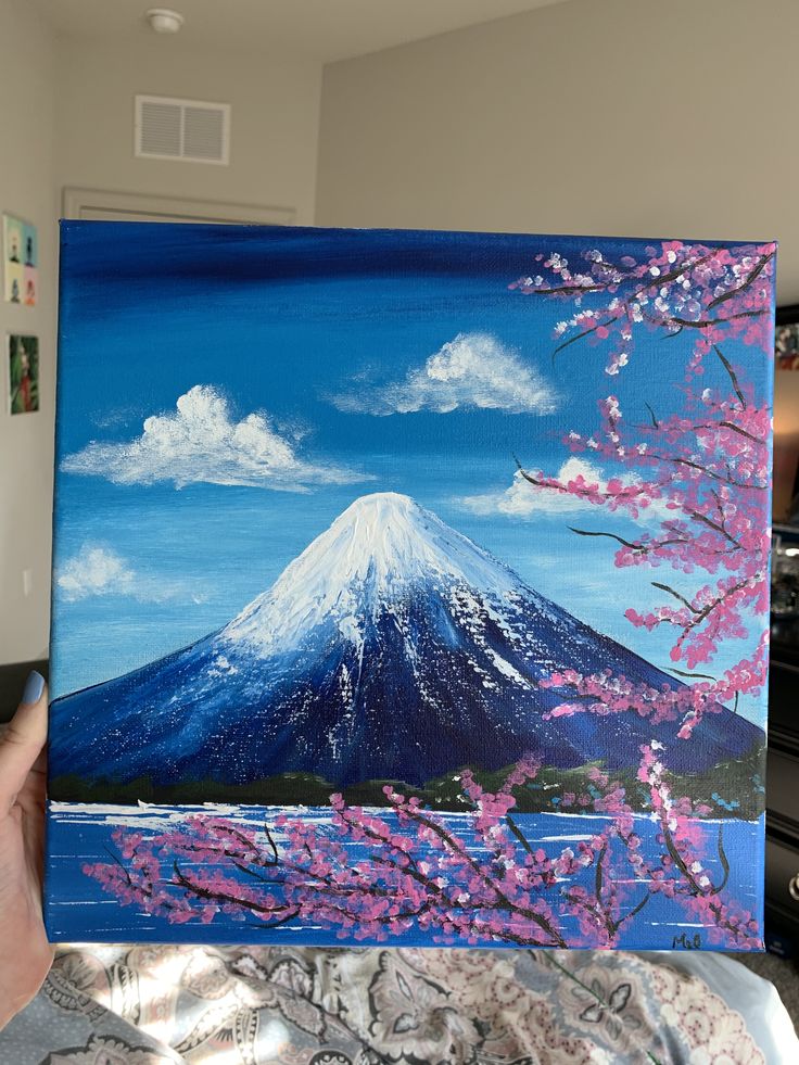 a person holding up a painting in front of a window with mountains and flowers on it