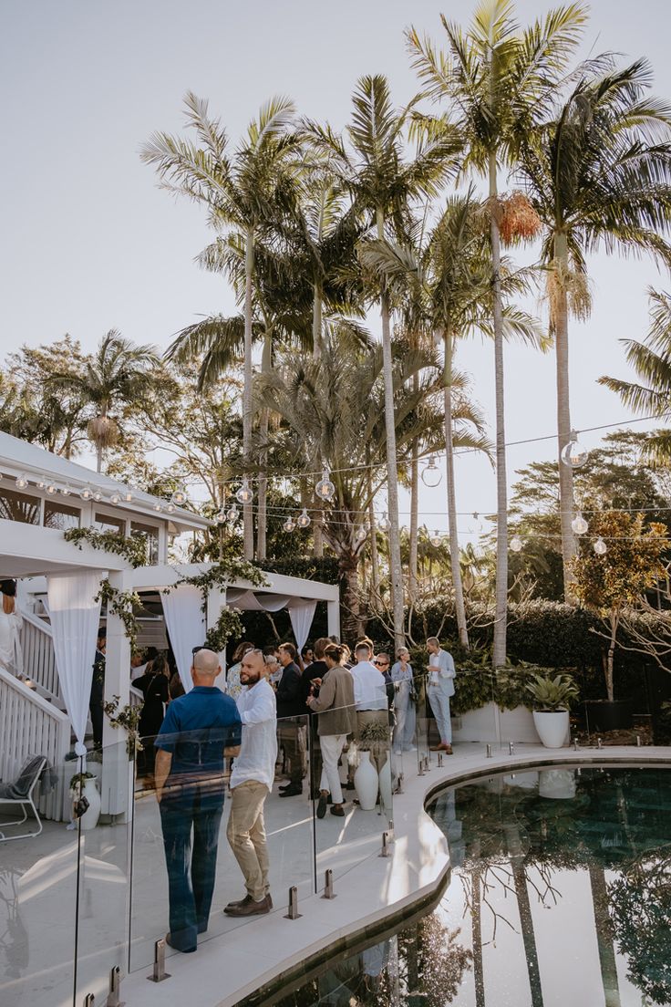 people are standing around in front of a pool with palm trees and white drapes