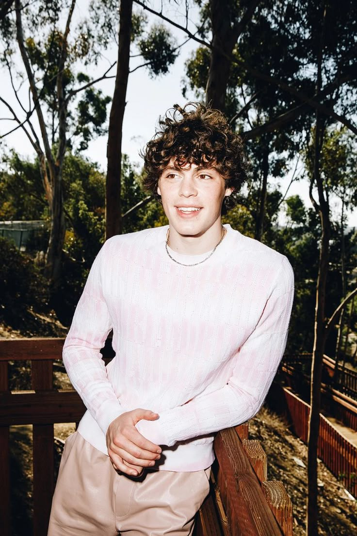 a young man standing on a wooden deck in front of some trees and bushes, wearing a pink sweater