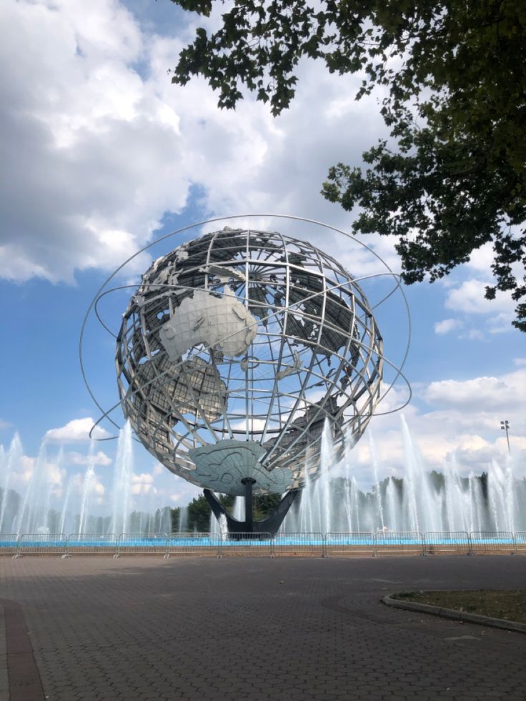 a large metal globe sitting in the middle of a park with water fountains behind it