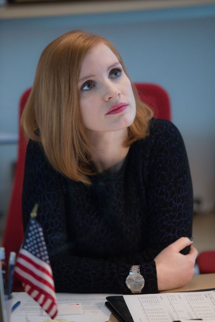 a woman sitting at a desk with an american flag in front of her and looking off to the side