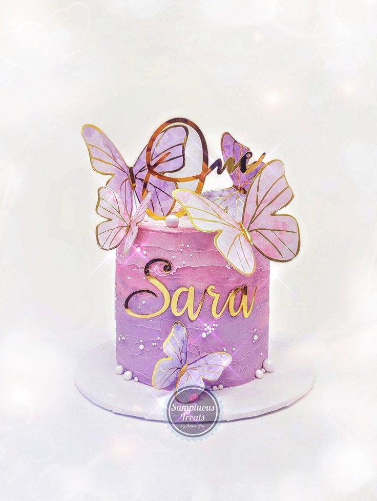 a pink cake with butterflies on top and the word saru spelled in gold letters