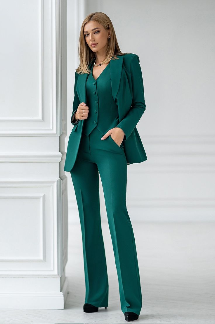 a woman in a green suit standing next to a white wall and looking at the camera