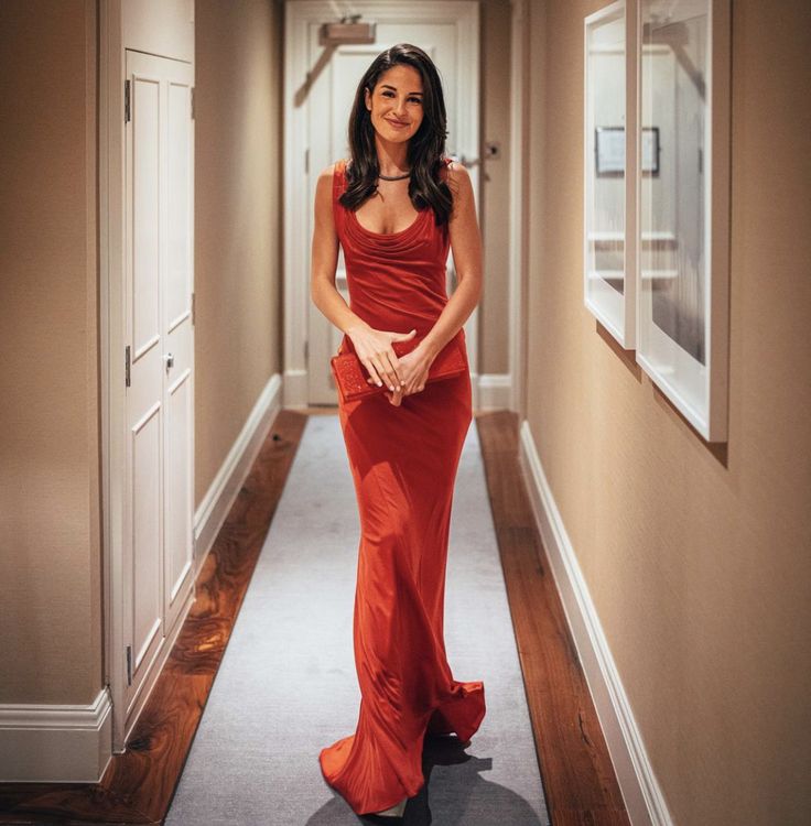 a woman in a long red dress is walking down the hallway with her hand on her hip