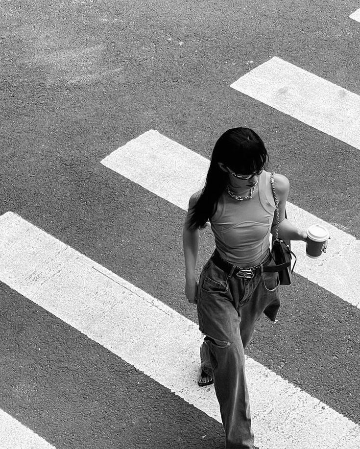 a woman walking across a cross walk holding a cup in one hand and a cell phone in the other