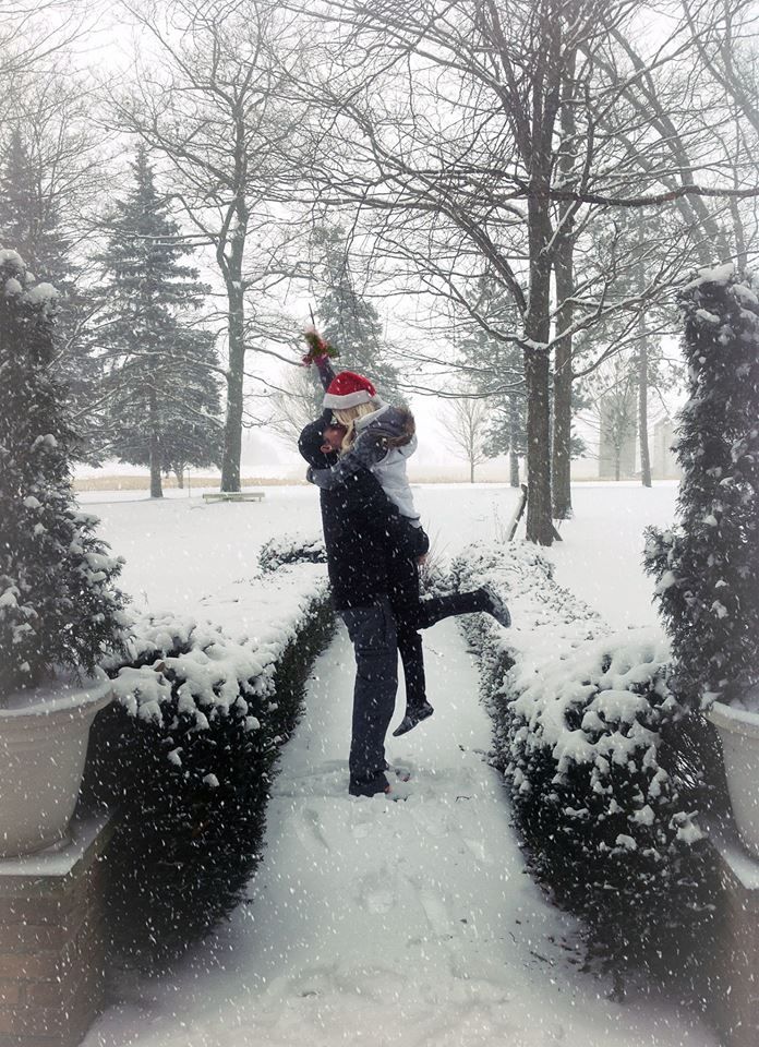 a man holding a child in the snow near some bushes and trees with snow falling on them