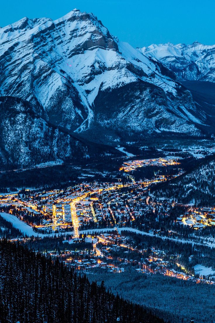 the city lights are lit up at night in front of snow covered mountains and trees