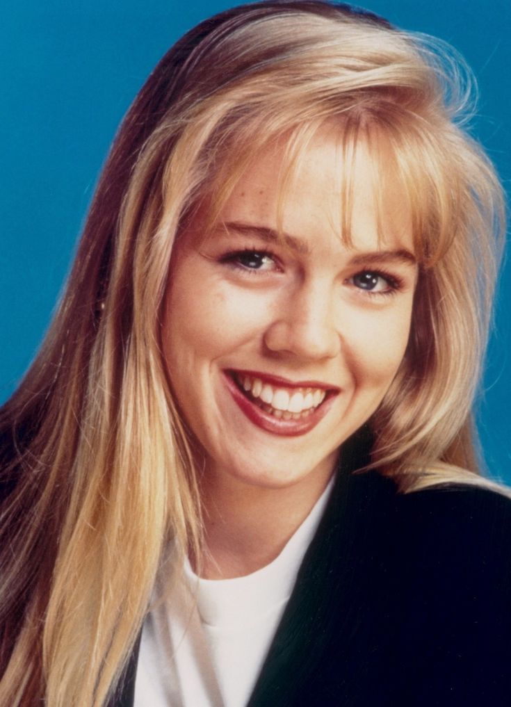 a woman with long blonde hair smiling at the camera while wearing a black blazer and white t - shirt