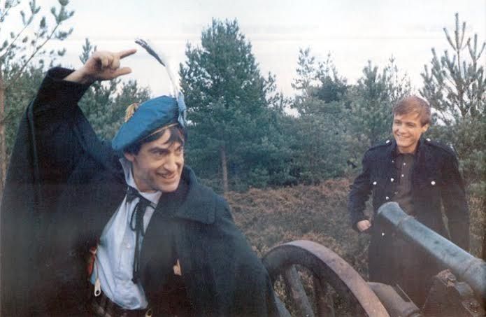 two young men dressed in medieval clothing standing next to each other near a fence and trees