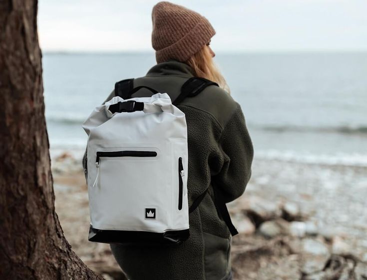 a person standing next to a tree looking at the ocean with a white backpack on it