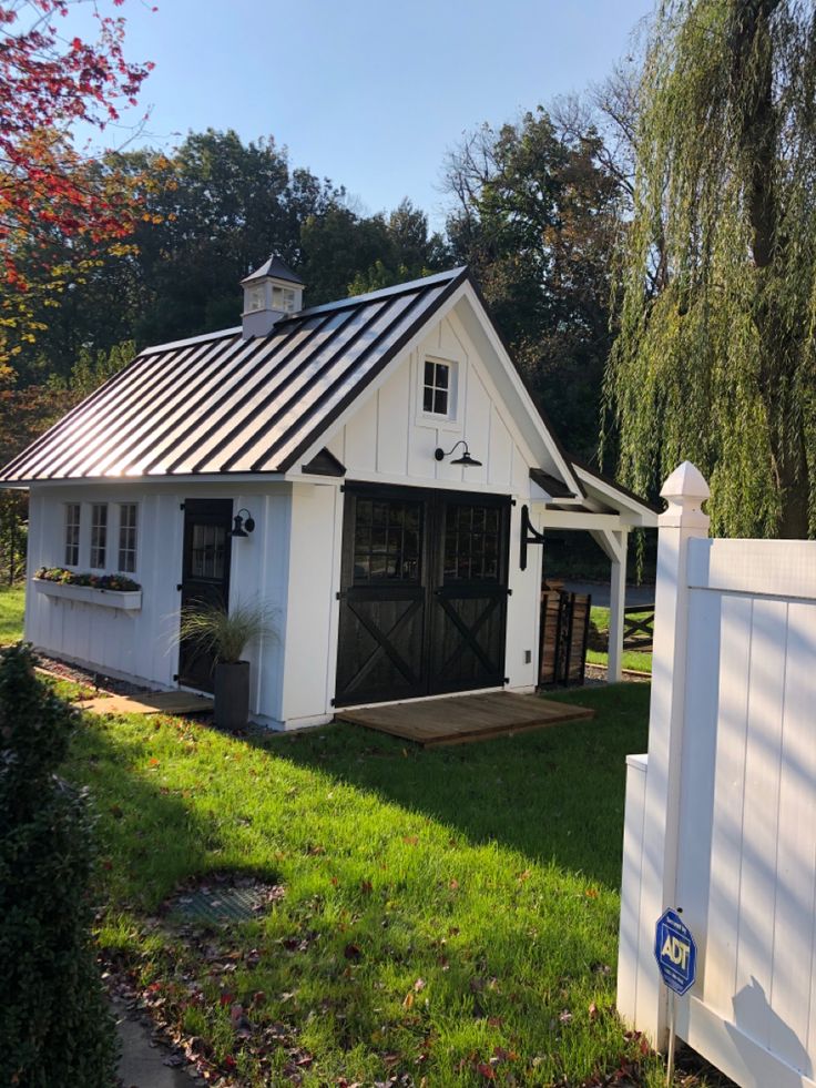 a small white barn with a black roof and door on the grass next to a fence