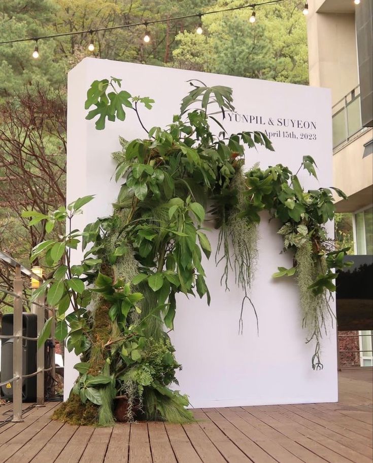 a sign with plants growing on it in front of a building that has trees and lights hanging from the roof