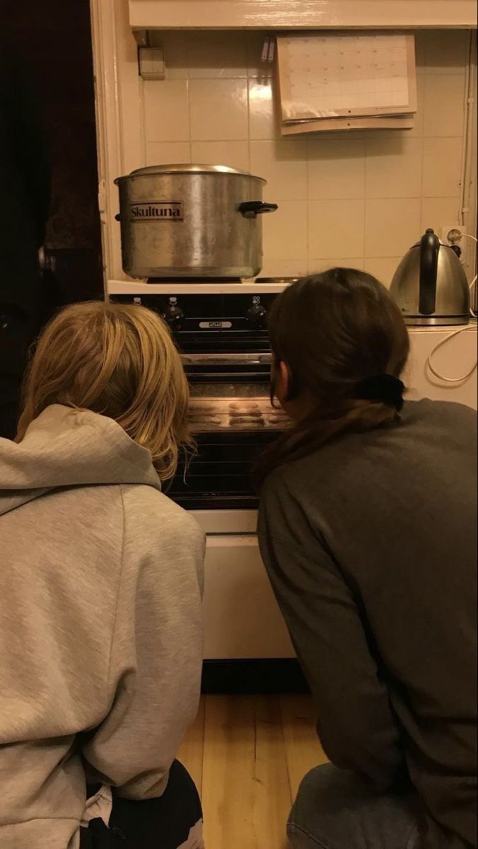 two people sitting in front of an oven with pots on the stove top and one person looking into it