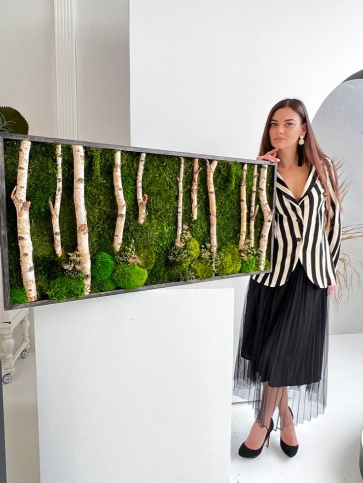 a woman standing in front of a moss wall with white trees and grass on it