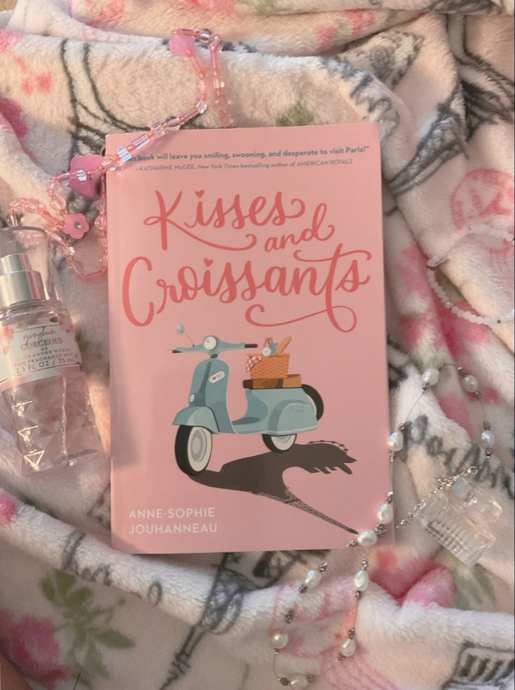 the book kisses and crisssants is laying on a blanket next to a bottle of cologne