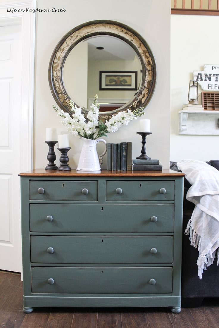 a dresser with flowers and books on it in front of a large round mirror above the dresser