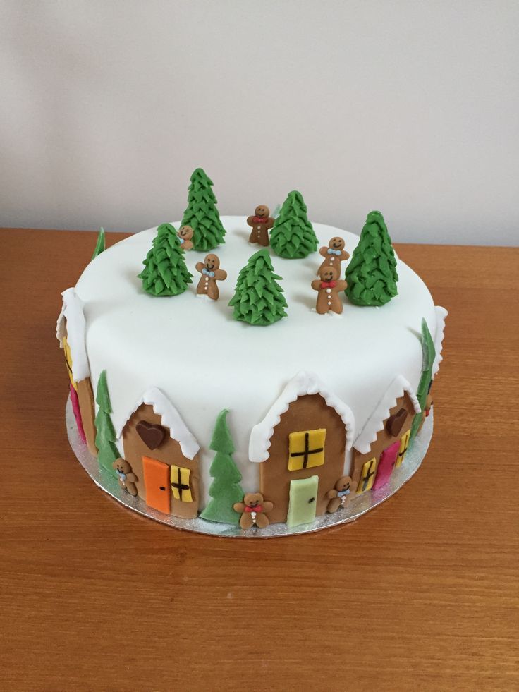 a white frosted christmas cake with trees and houses on the top is sitting on a wooden table