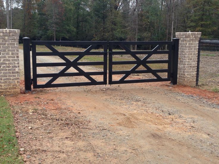 a black gate with brick pillars on the side of a dirt road in front of trees