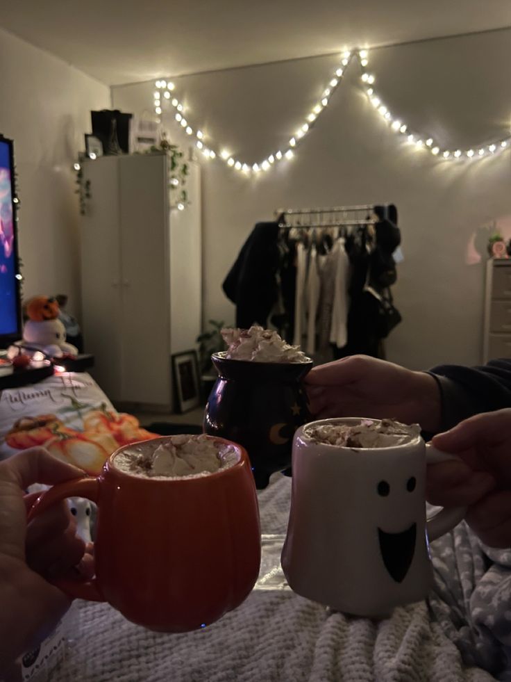 two people holding mugs in their hands on a bed with lights strung above them