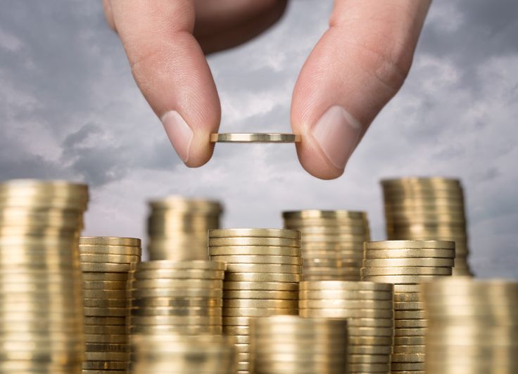 a hand is placing a coin on top of a pile of gold coins with clouds in the background