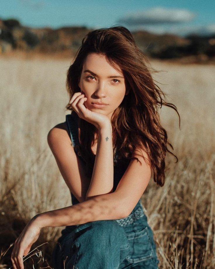 a beautiful young woman sitting in the middle of a field with her hand on her chin