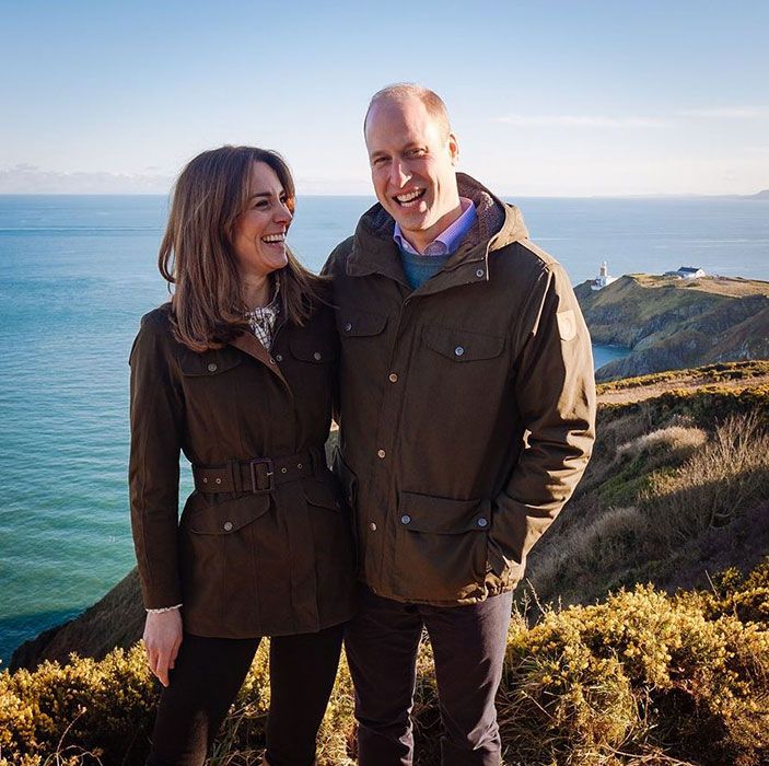 a man and woman standing on top of a hill next to the ocean with their arms around each other