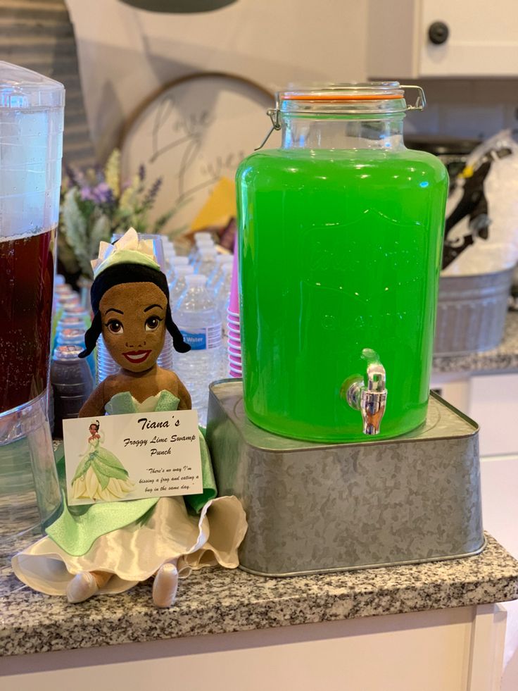 a plastic doll sitting on top of a counter next to a green liquid in a jar