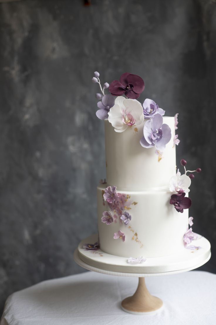 a three tiered white cake with purple and white flowers on the top, sitting on a table