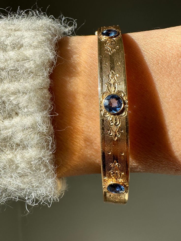 "Delicate and radiant, this one of a kind sapphire bangle would have made an unforgettable gift. Engraved on the rear of the bangle are the words \"Ultima from pop pop 1914\". The engraving on the rear matches the grace of the engraved design on the front. Both of these details are complimented by the use of faceted cornflower blue sapphires which have been bezel set. Estimated total sapphire weight 1.2 carats 14k gold 7mm thickness 60mm diameter 12.8 grams Carries engraving \"Ultima from pop po Luxury Gemstone Cuff Bracelet For Anniversary, Luxury Yellow Gold Cuff Bracelet With Gemstone, Luxury Blue Gemstone Bangle, Luxury Yellow Gold Collectible Bangle, Luxury Sapphire Bangle Bracelet, Luxury Sapphire Bangle Bracelets, Luxury Collectible Yellow Gold Bangle, Blue Luxury Bangle For Formal Occasions, Luxury Oval Gemstone Bangle