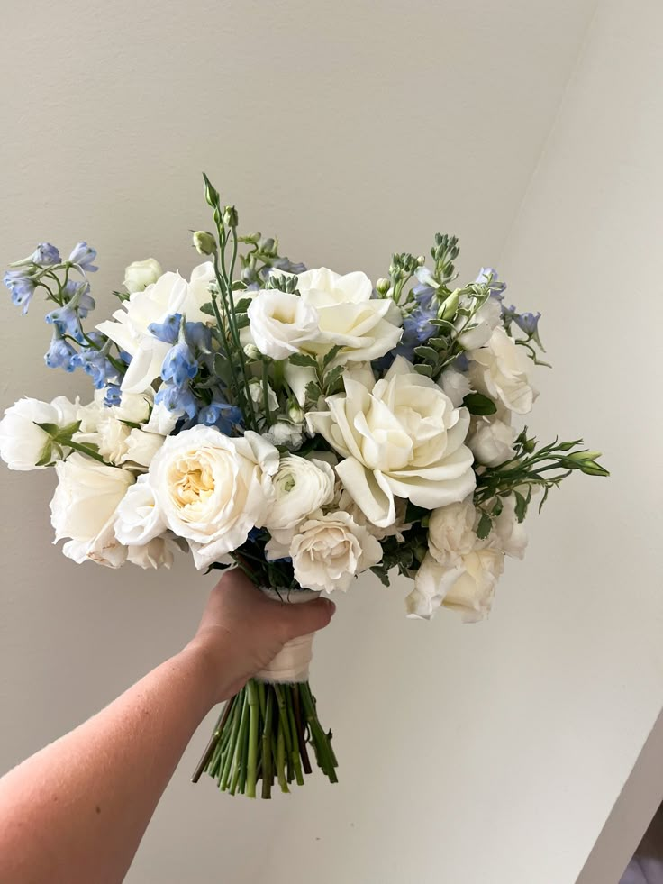 a person holding a bouquet of white and blue flowers in their left hand, against a wall