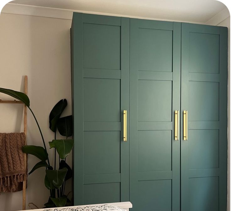 a room with green cupboards and a white bed in it's corner, next to a large potted plant