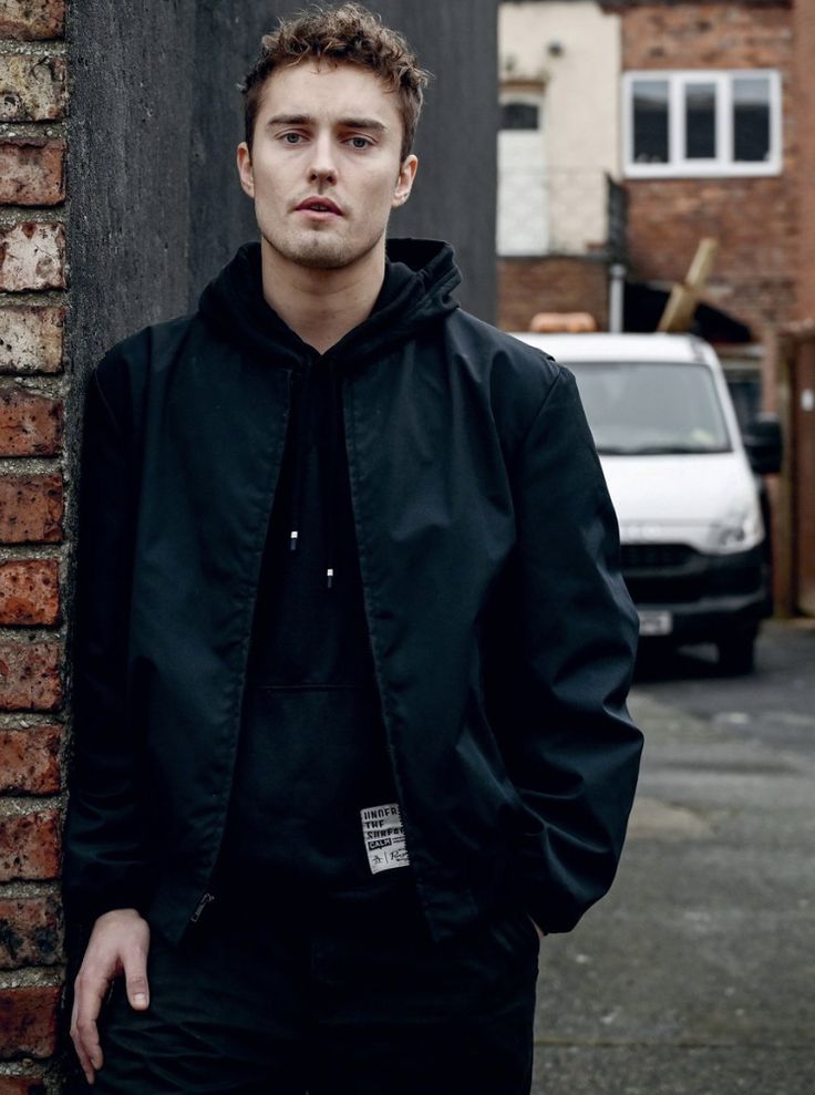 a young man standing next to a brick wall wearing a black jacket and dark pants