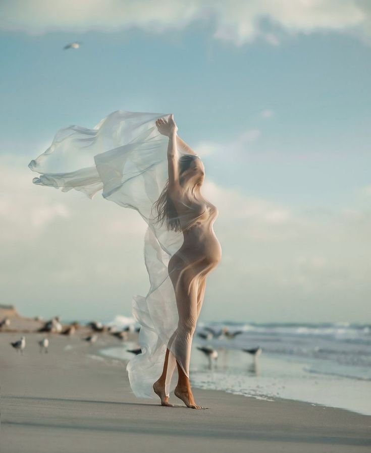 a nude woman standing on top of a sandy beach next to the ocean and seagulls