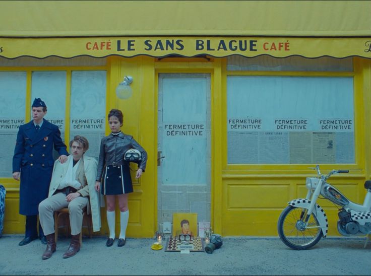 three people standing in front of a yellow building with two motorcycles parked outside the door