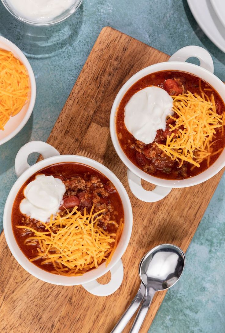 two white bowls filled with chili and cheese on top of a wooden cutting board next to silverware