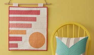 a yellow chair next to a wall hanging with a colorful pillow on top of it