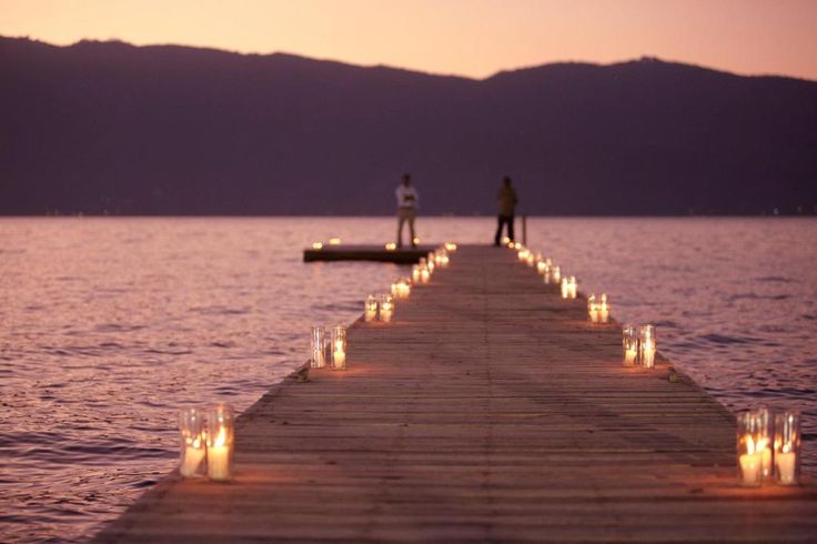 two people standing on the end of a dock with candles lit up in front of them