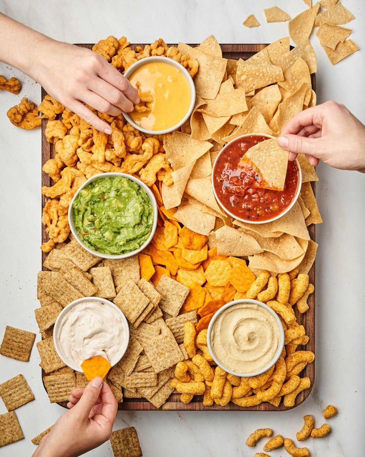 two hands reaching for chips and dips on a tray filled with tortilla chips