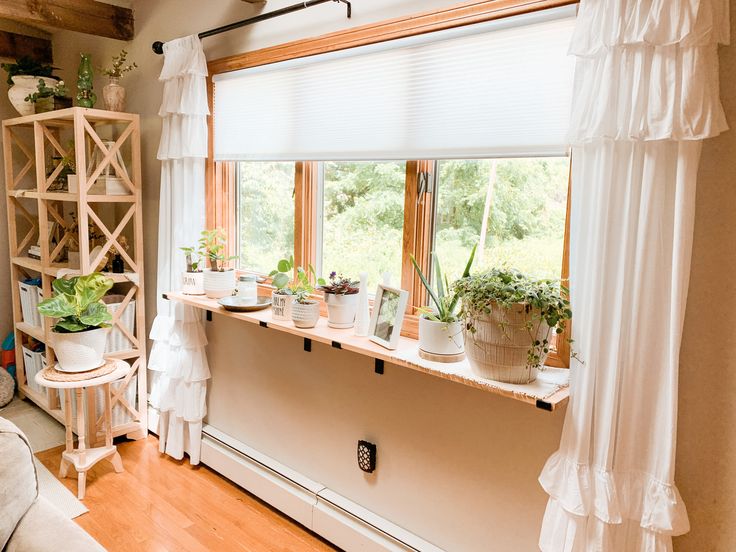 a living room filled with lots of plants next to a large window covered in white drapes