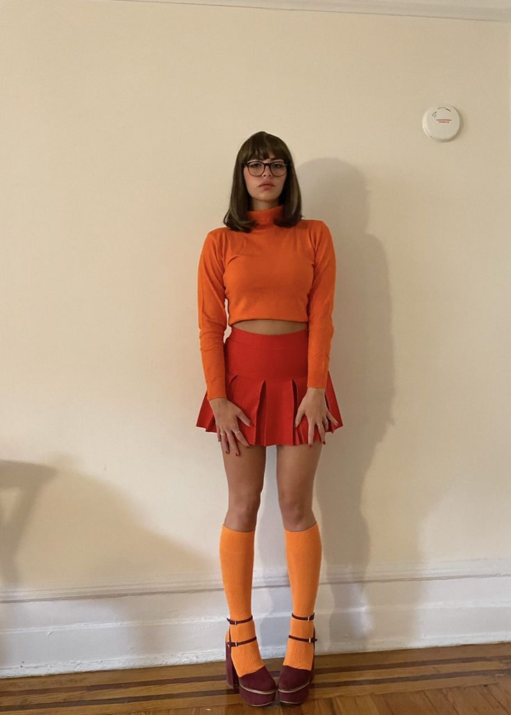 a woman standing in front of a white wall wearing an orange top and red skirt