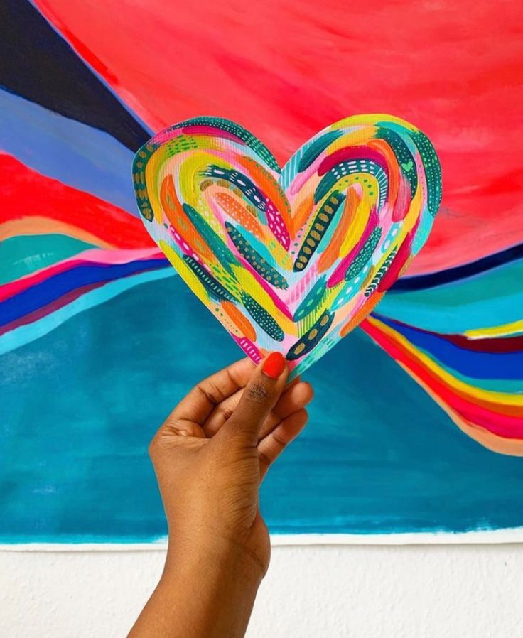 a person holding up a heart shaped paper piece in front of a colorful wall painting