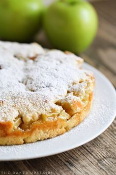 an apple pie on a white plate with two green apples in the background