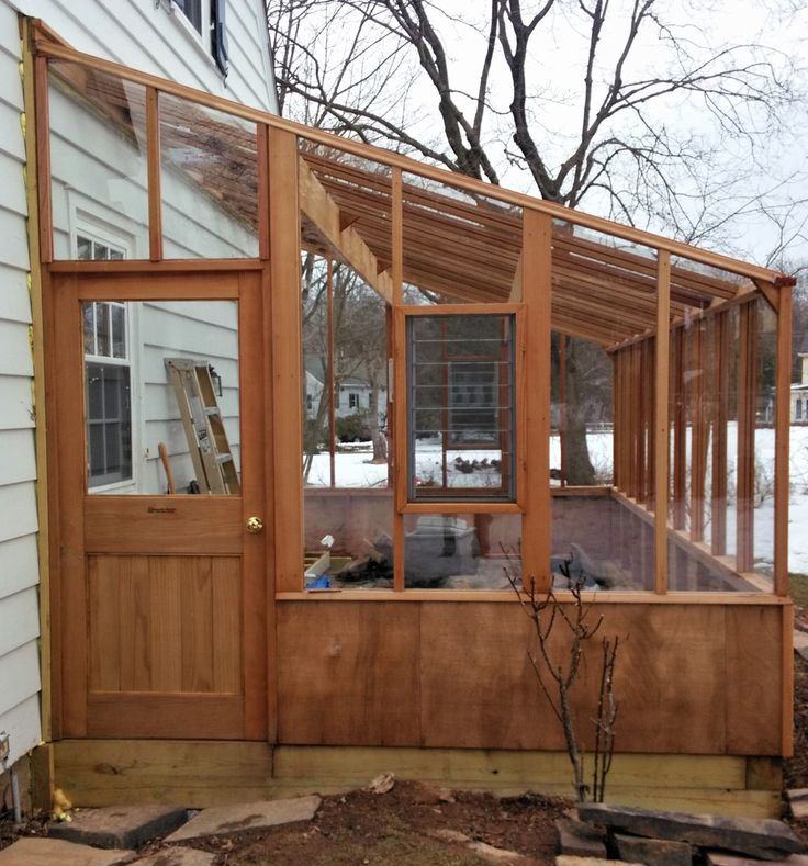 a small wooden greenhouse in front of a house with snow on the ground and bare trees