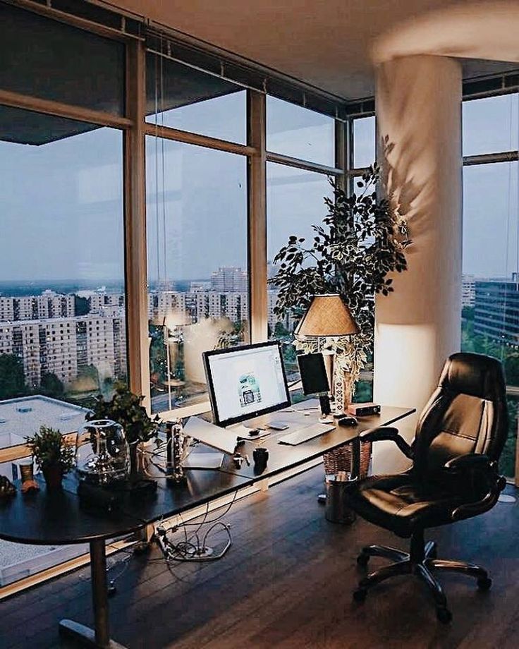 a desk with a computer, lamp and plant in front of large windows overlooking the city