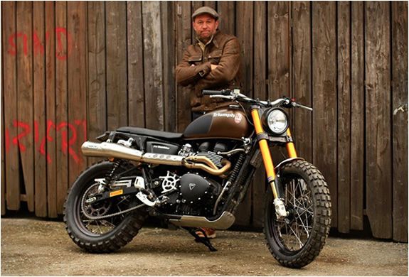 a man standing next to a motorcycle parked in front of a wooden wall with graffiti on it