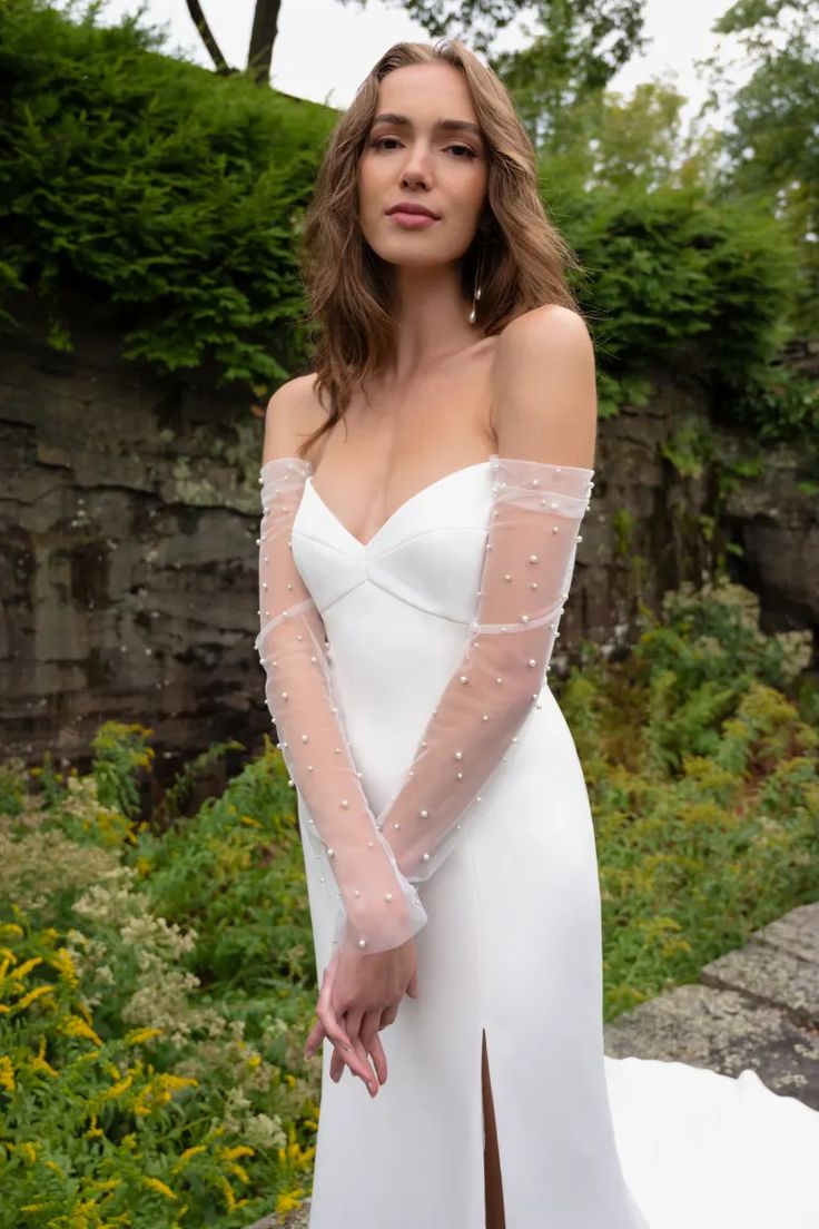 a woman wearing a white dress with long sleeves and pearls on the shoulder, standing in front of flowers