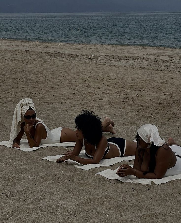 three women in bathing suits laying on the sand at the beach with towels around them